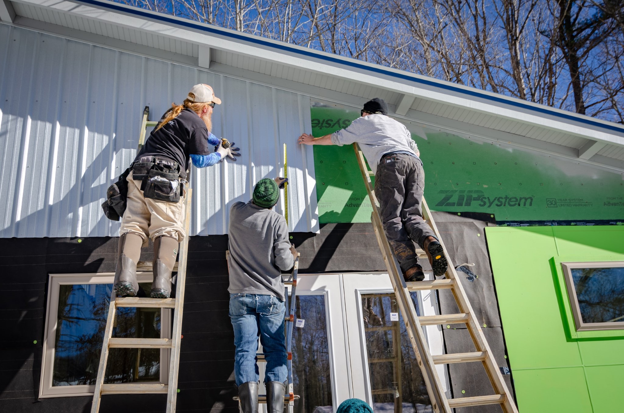 Tre personer står på stegar och monterar panel på ett hus.