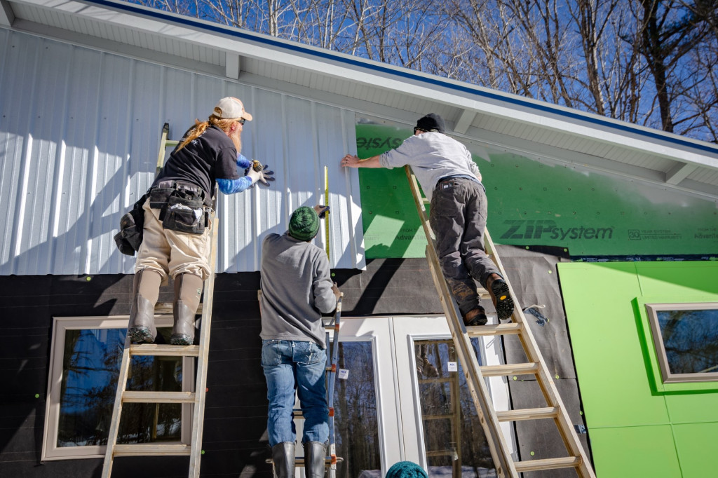 Tre personer står på stegar och monterar panel på ett hus.