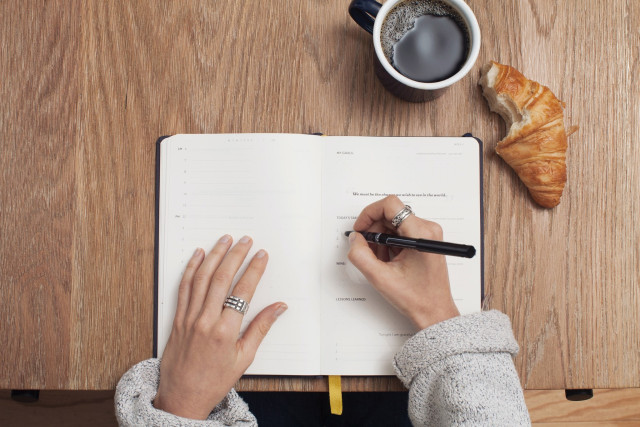 Person som antecknar i en öppen bok, med en kopp kaffe och en croissant bredvid på bordet.